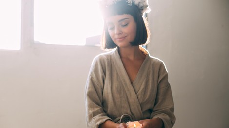 Tender and sensual photo of young natural beautiful woman in cotton linen organic bath robe holding melted wax candle, prepares for relaxation or spa at cosy home